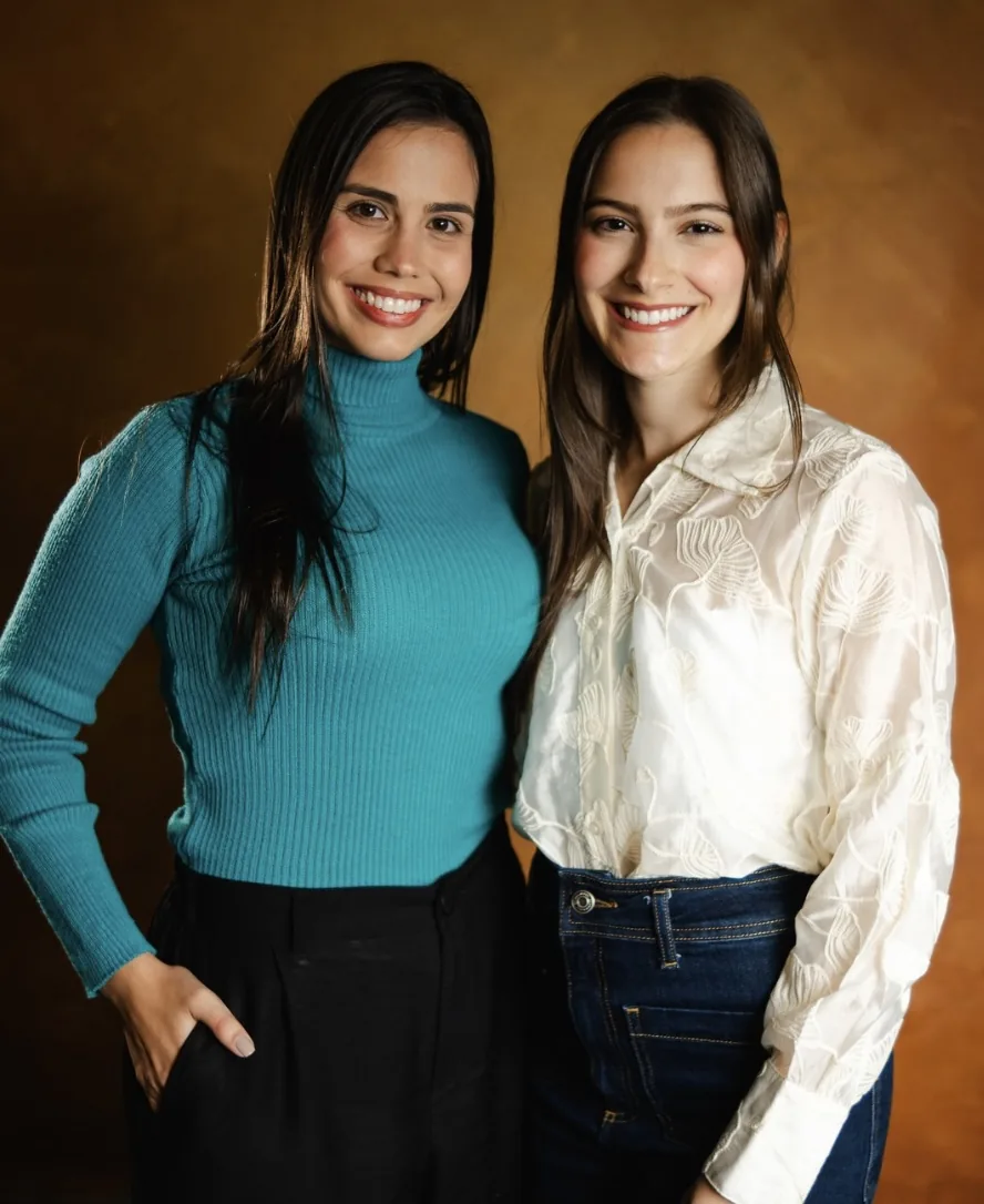 As duas sócias do escritório, lado a lado e sorrindo, diante de um fundo em tons de dourado